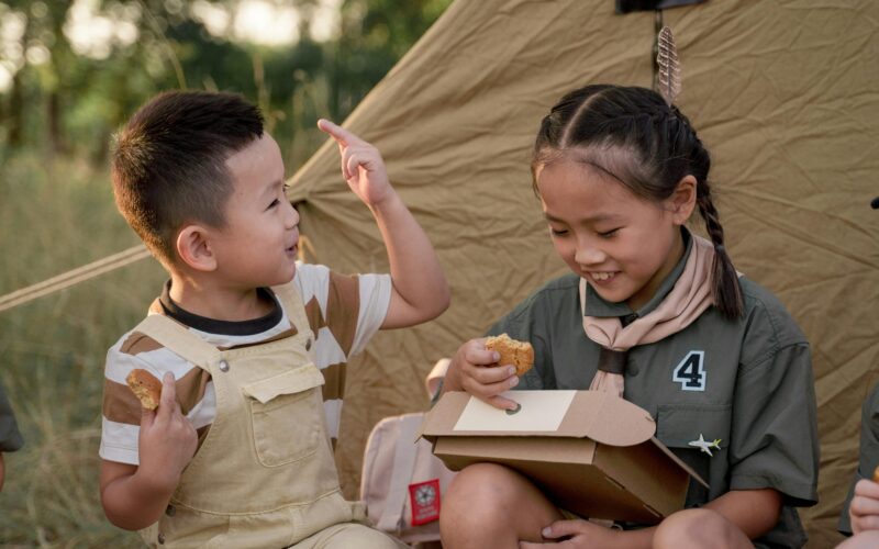 Two Asian kids (a boy and a girl) eating beside a camping tent