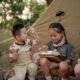 Two Asian kids (a boy and a girl) eating beside a camping tent