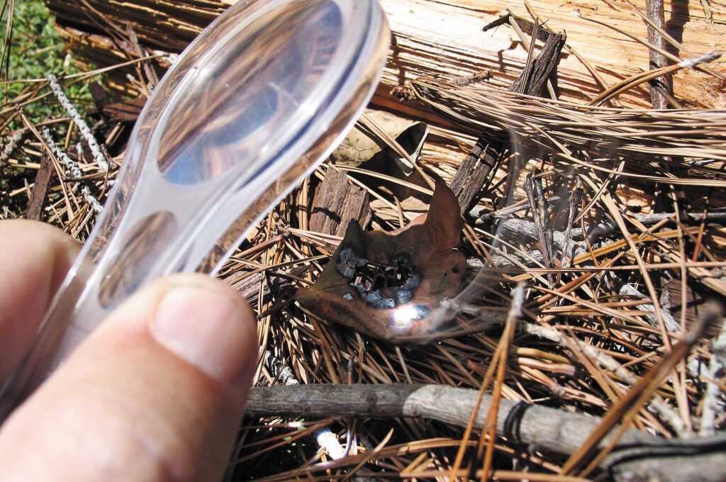 A lens is focused on sunlight to be used to strike fire on on some grass as a technique on  how to start a fire without matches or lighter